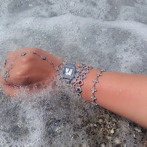Plusieurs bracelets, bagues et une montre + une chaine de main argentés en acier inoxydable portée par une femme sur une plage dans l'eau 